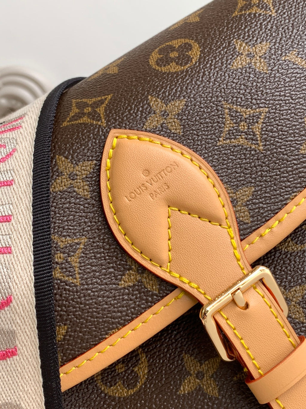Close-up of monogram canvas flap with tan leather trim, yellow stitching, gold hardware