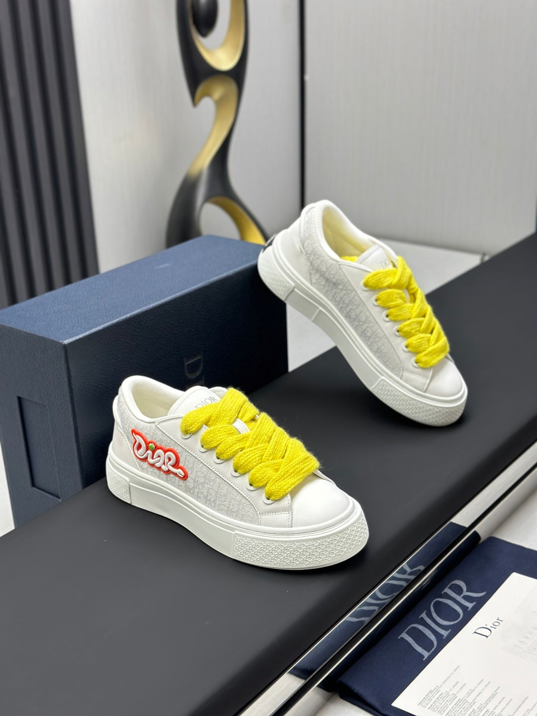 White low-top sneakers with yellow laces and red logo, angled view on shoebox.