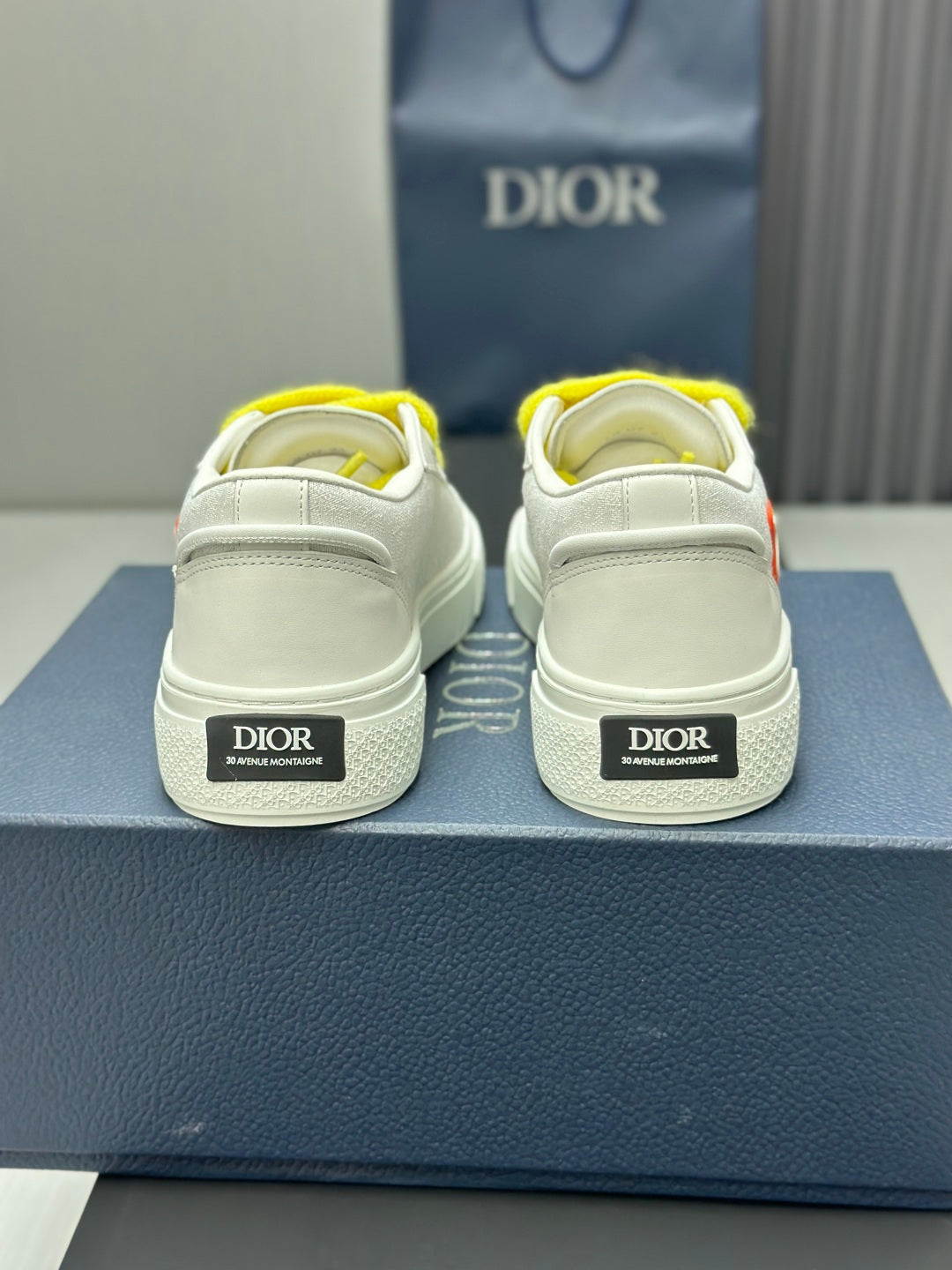White low-top sneakers with yellow laces, rear view with Dior logo heel labels.
