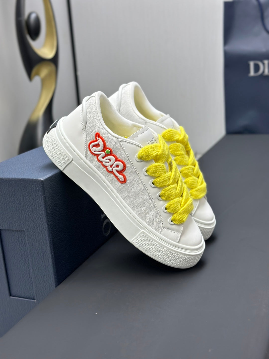 White low-top sneaker with yellow laces and red logo embroidery, close-up on box.