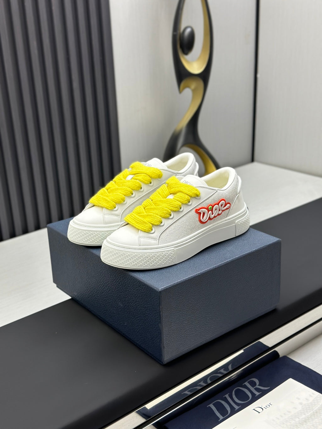 White low-top sneakers with yellow laces and red logo detail, on blue shoebox.
