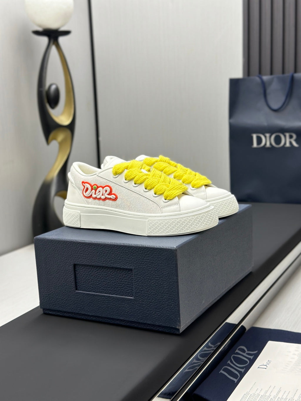 White low-top sneakers with yellow laces and red logo patch, side view on box.