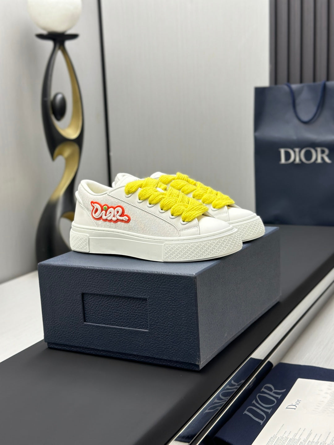White low-top sneakers with yellow laces and red logo patch, side view on box.