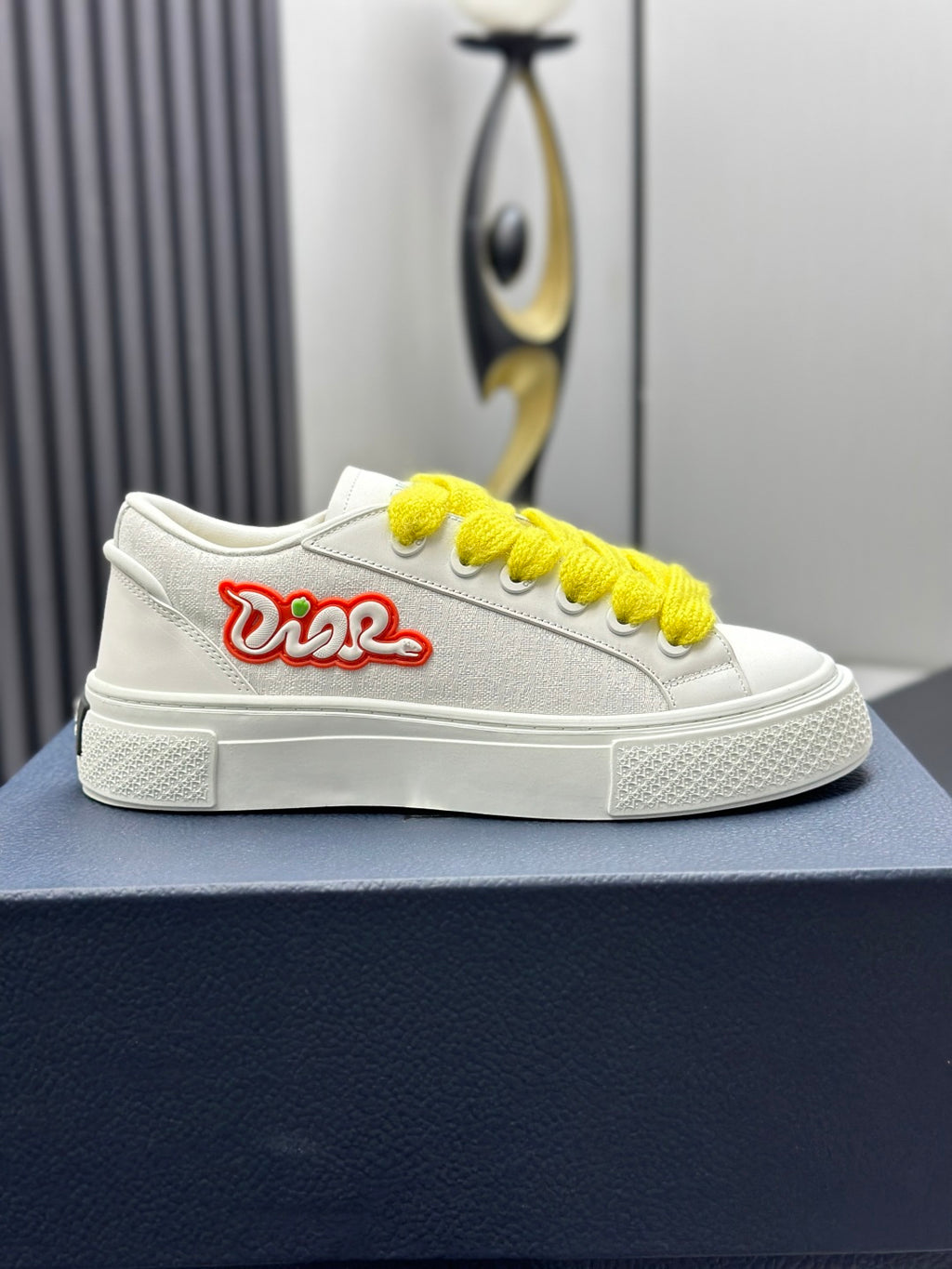 White low-top sneakers with yellow laces and red logo, front view on blue shoebox.