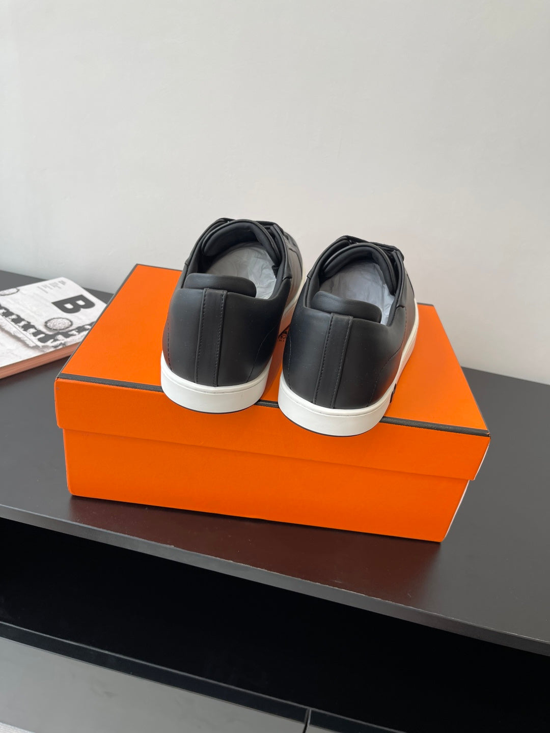 Rear view of black low-top sneakers with lace-up tops and white rubber sole, on orange box.