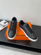 Two black leather low-top sneakers with lace-up front and white sole, side-by-side on orange box.