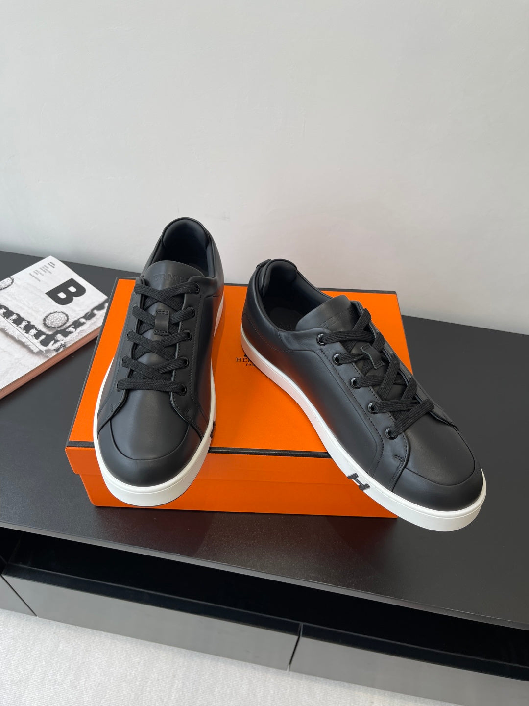 Two black leather low-top sneakers with lace-up front and white sole, side-by-side on orange box.