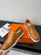 Brown leather slip-on loafers with white soles; side view pair on orange box.