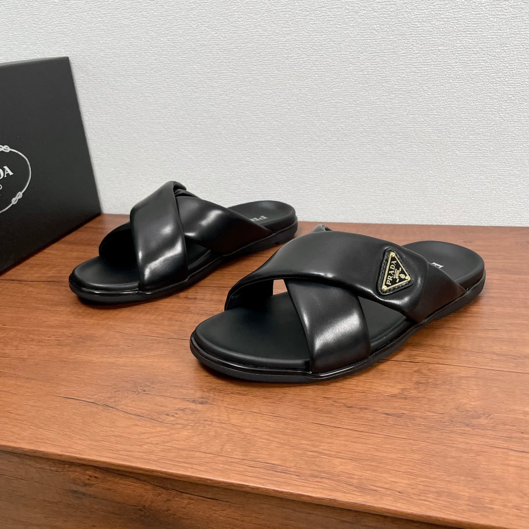 Black leather slide sandals with cross straps and gold logo plaque on upper