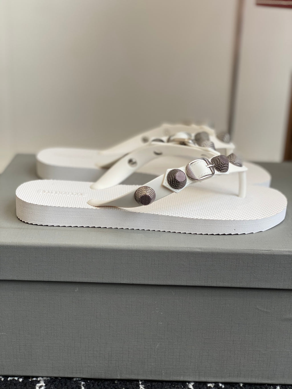 White thong sandals on box, featuring metallic buckle and round stud detail