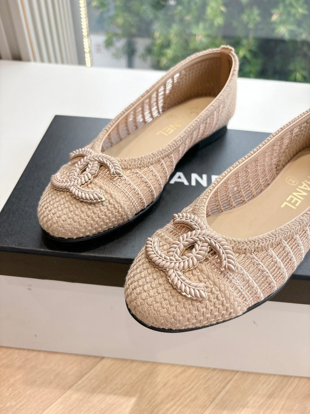 Beige woven ballet flats with braided bow detail; CHANEL logo on box behind shoes.
