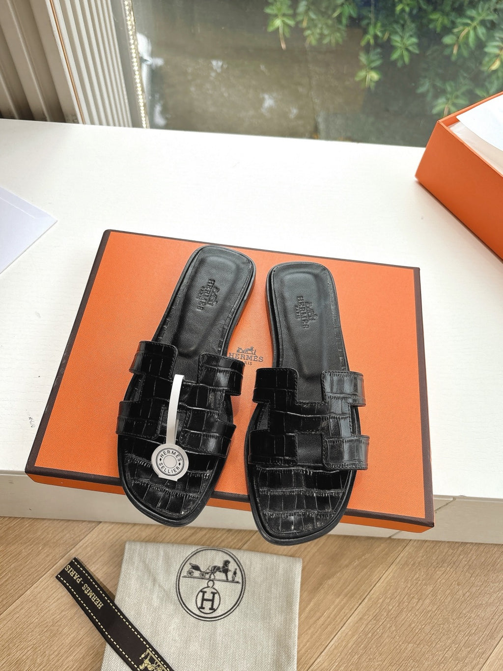 Black slide sandals with buckle straps and circular logo charm, displayed on orange box