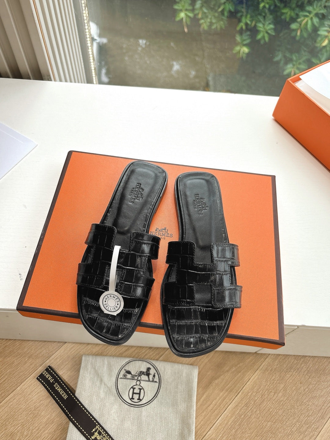 Black slide sandals with buckle straps and circular logo charm, displayed on orange box