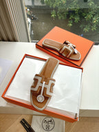 Tan-and-brown leather slide sandals on orange box, geometric strap and logo emblem