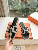 Hermes slide sandals, black with white H-style straps, held above orange shoebox.