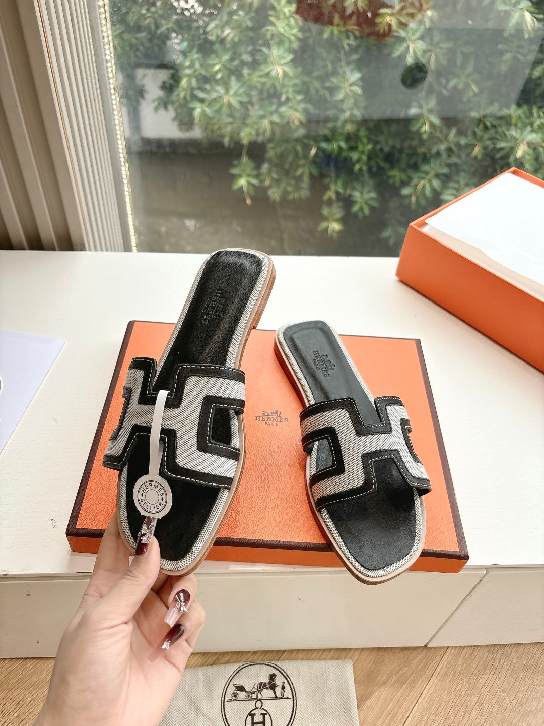 Hermes slide sandals, black with white H-style straps, held above orange shoebox.