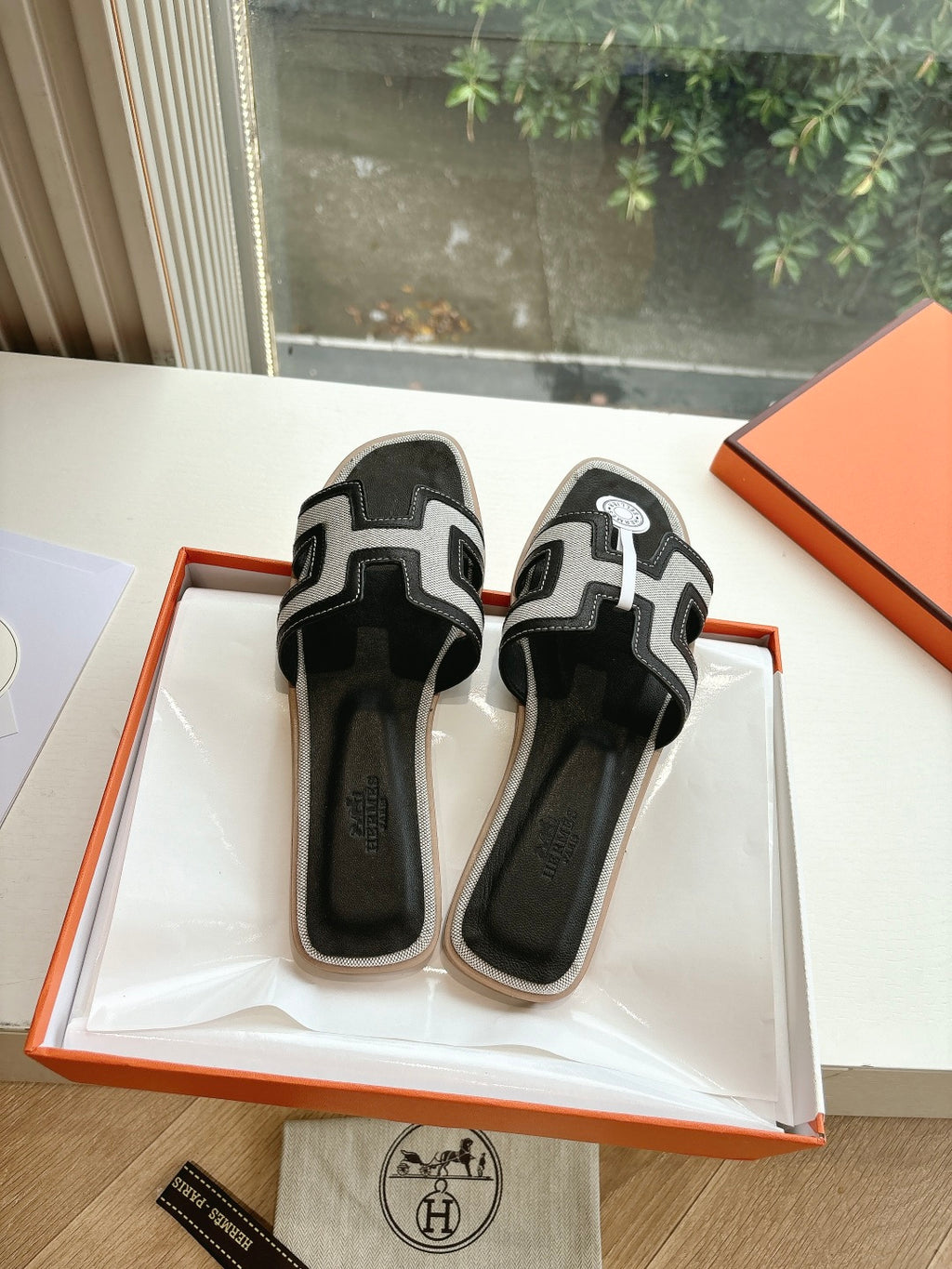 Pair of Hermes slide sandals in black with white H-style straps, open shoebox.