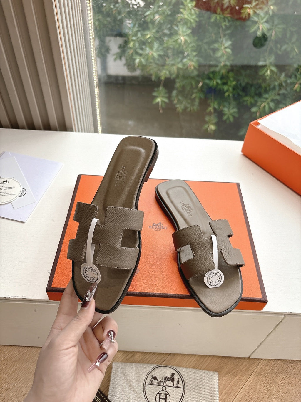 Dark taupe leather slide sandals held in hand, showing front strap and logo medallion