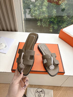 Dark taupe leather slide sandals held in hand, showing front strap and logo medallion