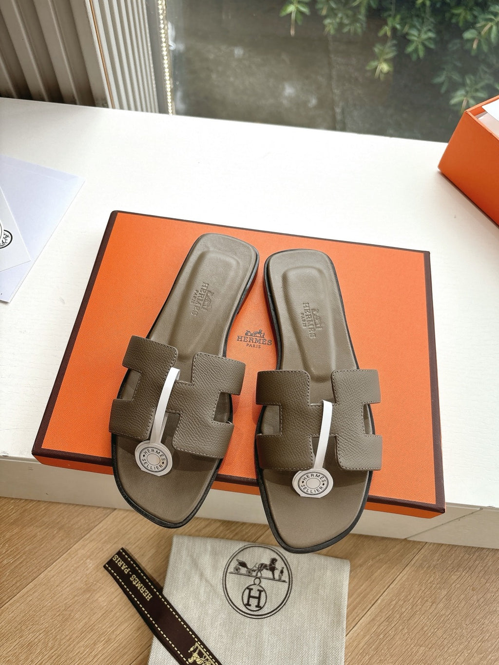 Dark taupe leather slide sandals on orange box lid; front straps with round logo
