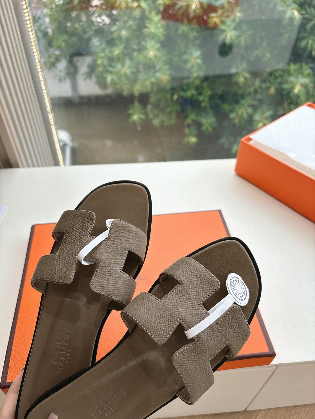 Dark taupe leather slide sandals with front strap and round logo emblem, angled view