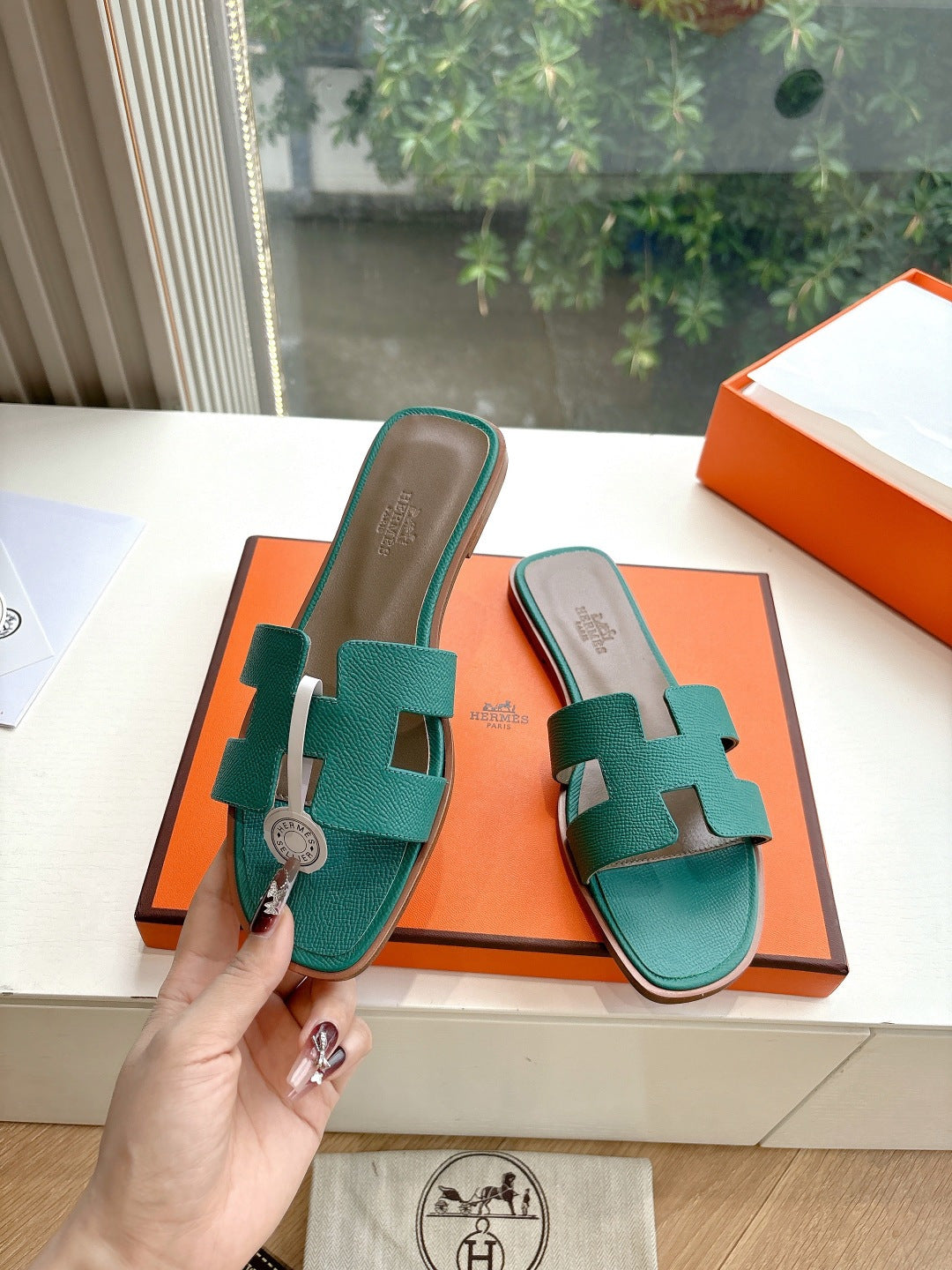Hermes ladies sandals, teal green H-strap slides held in hand above orange box