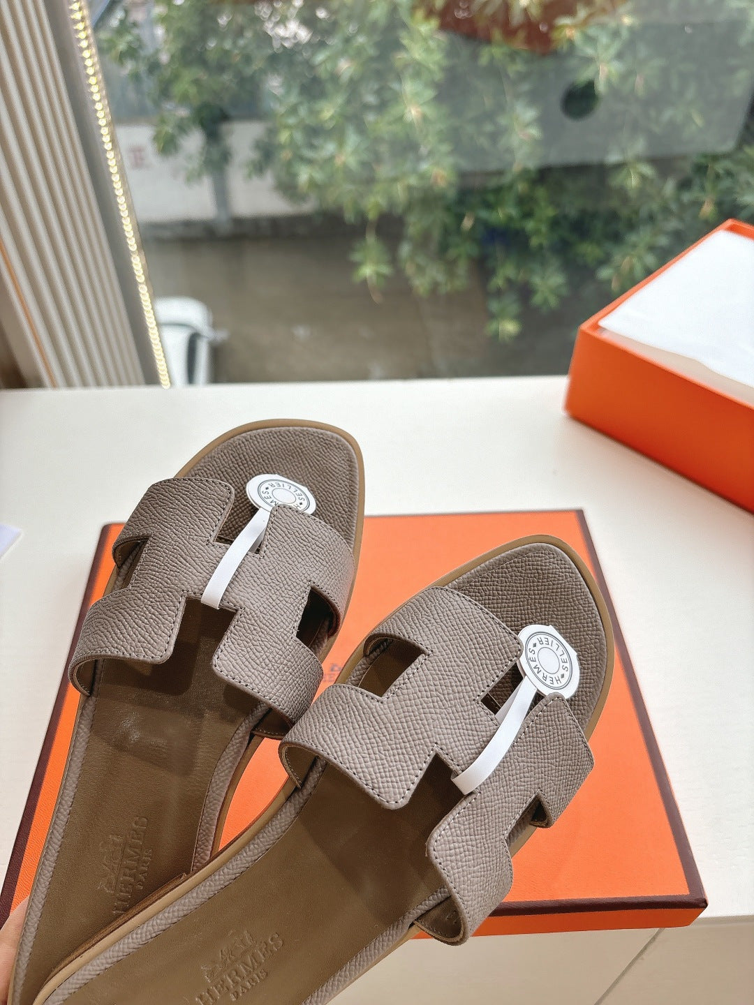 Taupe Hermès slide sandals close-up, adjustable straps with round logo medallions