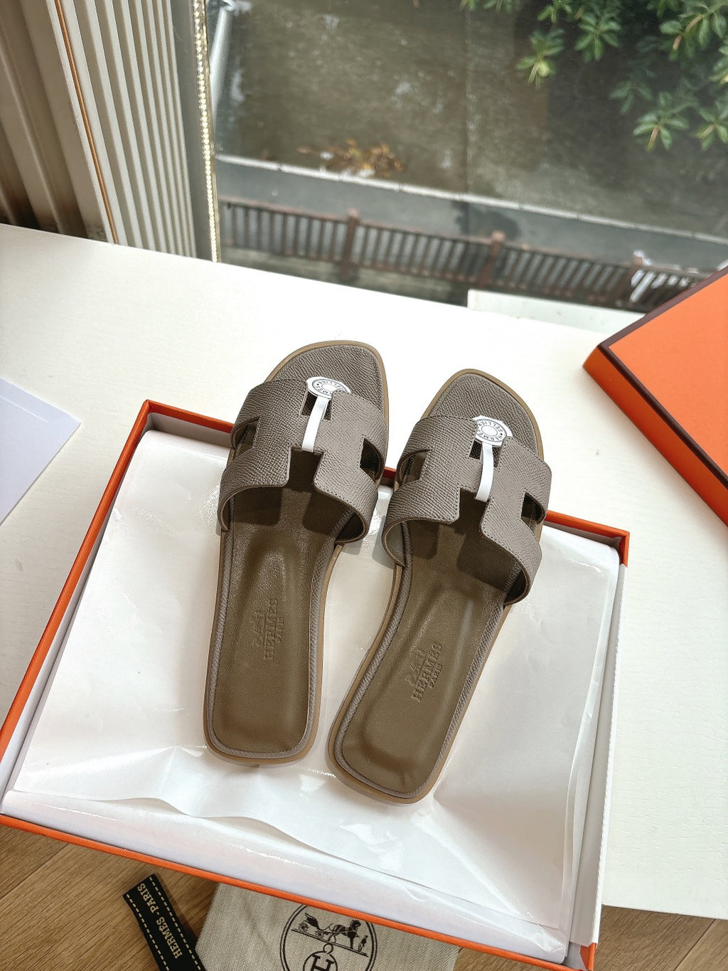 Taupe Hermès slide sandals pair view with white strap details and round logo medallion