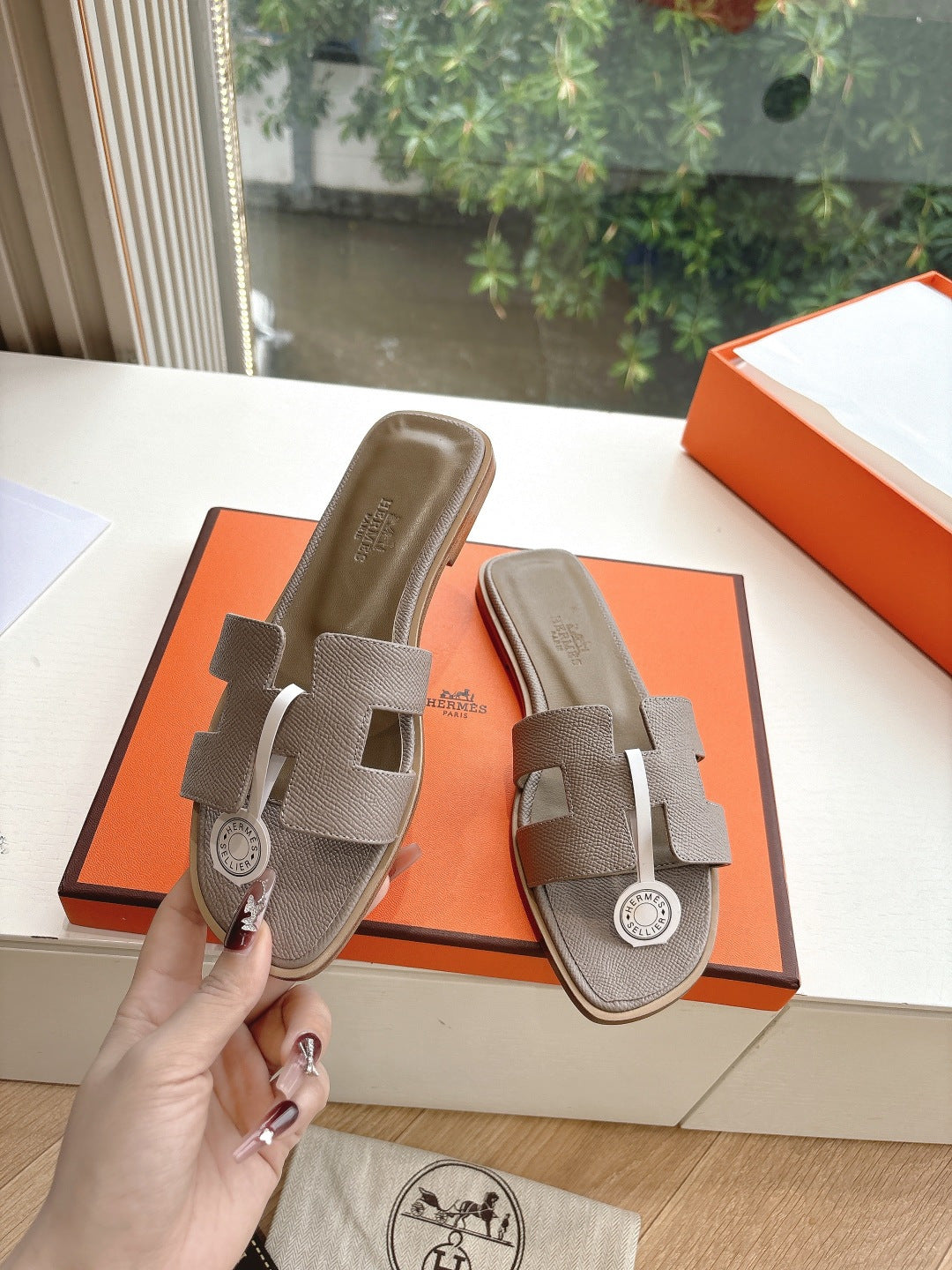 Taupe Hermès slide sandals held in hand, showing strap and round logo medallion