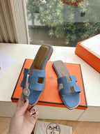 Blue Hermes slide sandals with H cutout held in hand above orange shoe box