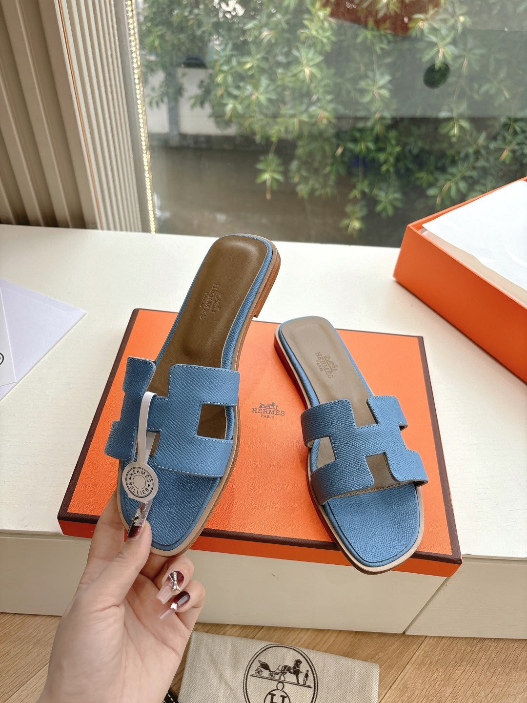 Blue Hermes slide sandals with H cutout held in hand above orange shoe box