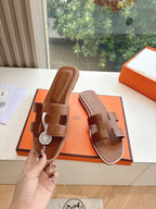Hermes leather slide sandals held in hand, brown with logo medallion strap