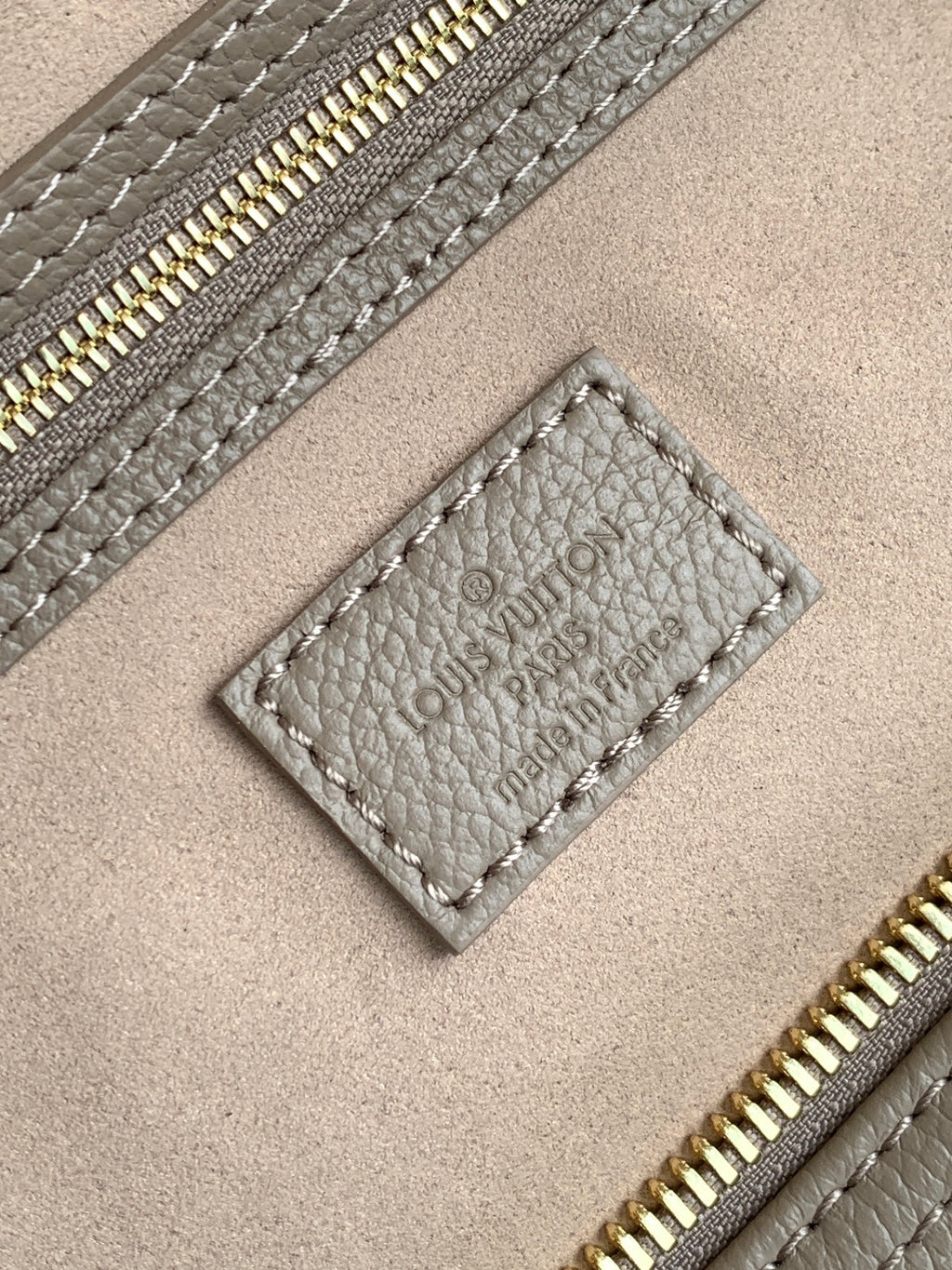 Louis Vuitton bag close-up: taupe leather label and gold zipper on textured grain