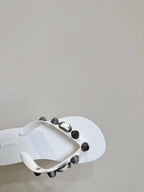 White thong sandals with studded oval hardware on the strap, angled side close-up.