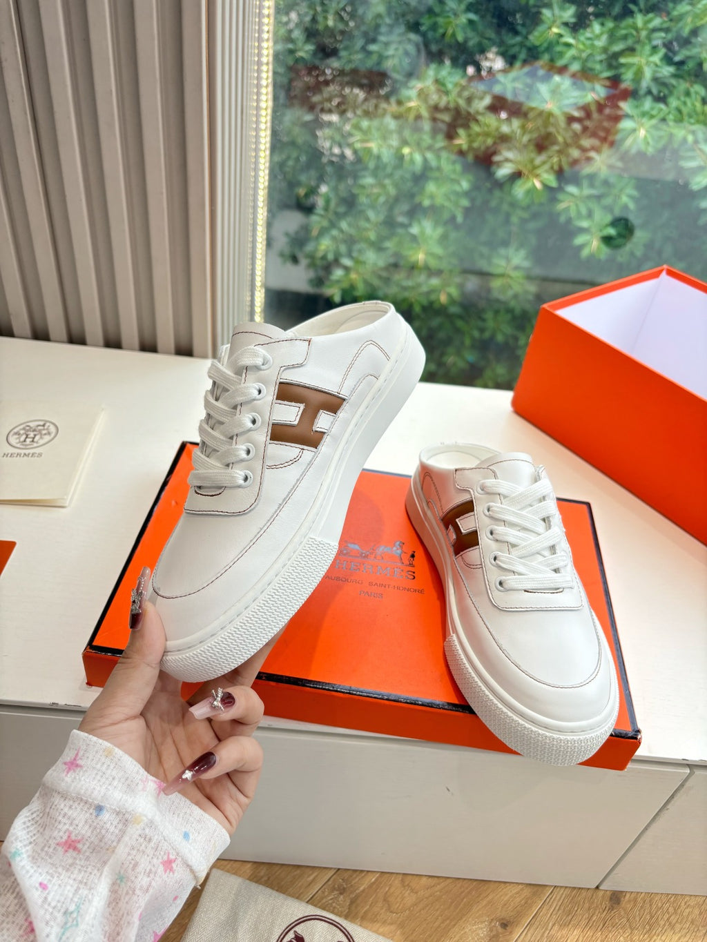 White Hermes sneakers for ladies, one held up with brown H side detail