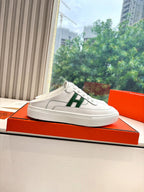 White slide-on sneakers with green H logo panel on side, on orange box