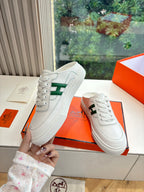 White low-top sneakers with green H logo, held in hand near orange box