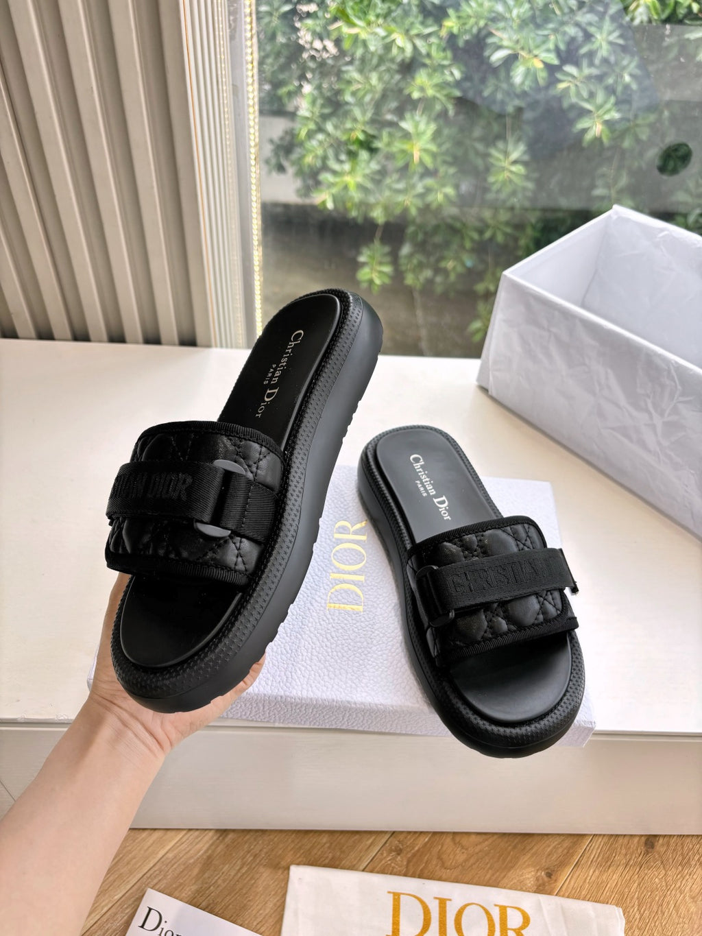 Black slide sandals on box with textured strap and cushioned sole; one shoe held up