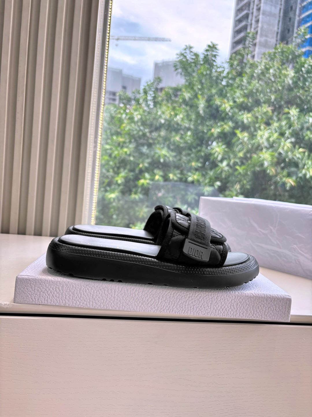 Black slide sandal with textured strap, shown in profile on top of the shoebox