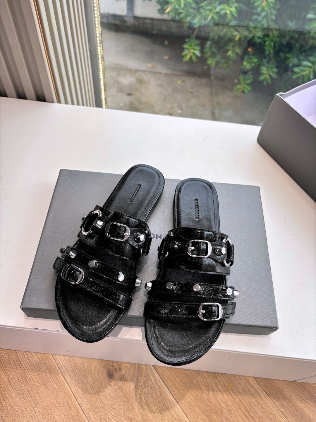 Black slide sandals with silver buckles on double straps, pair on box