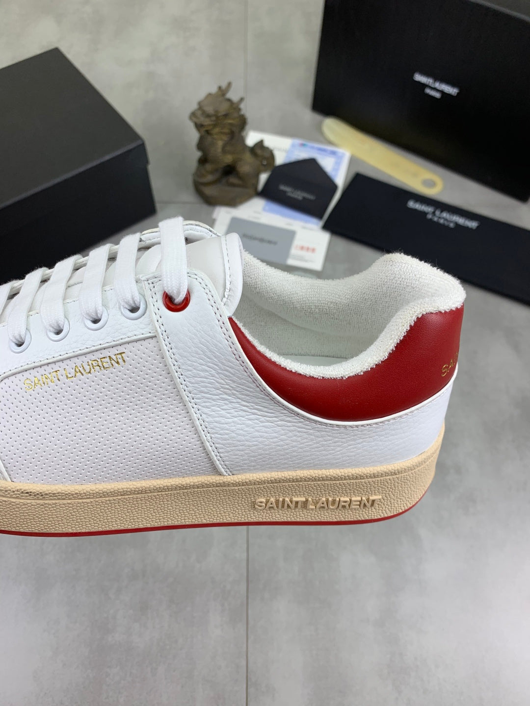 White sneaker with red heel stripe and stitched “Saint Laurent” logo on side.