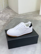 White low-top sneaker on a black box, black heel accent and gum sole.