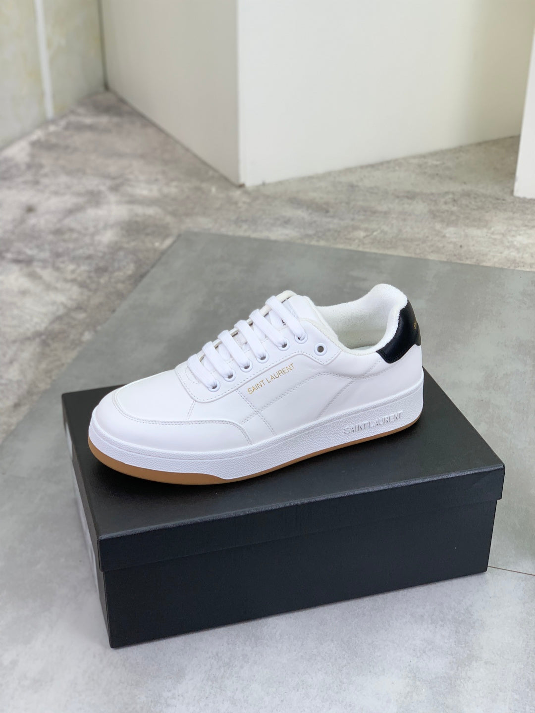 White low-top sneaker on a black box, black heel accent and gum sole.