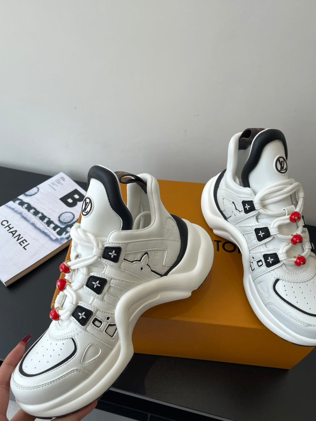 White athletic sneakers with black details, embossed logos, red pom-pom lace beads