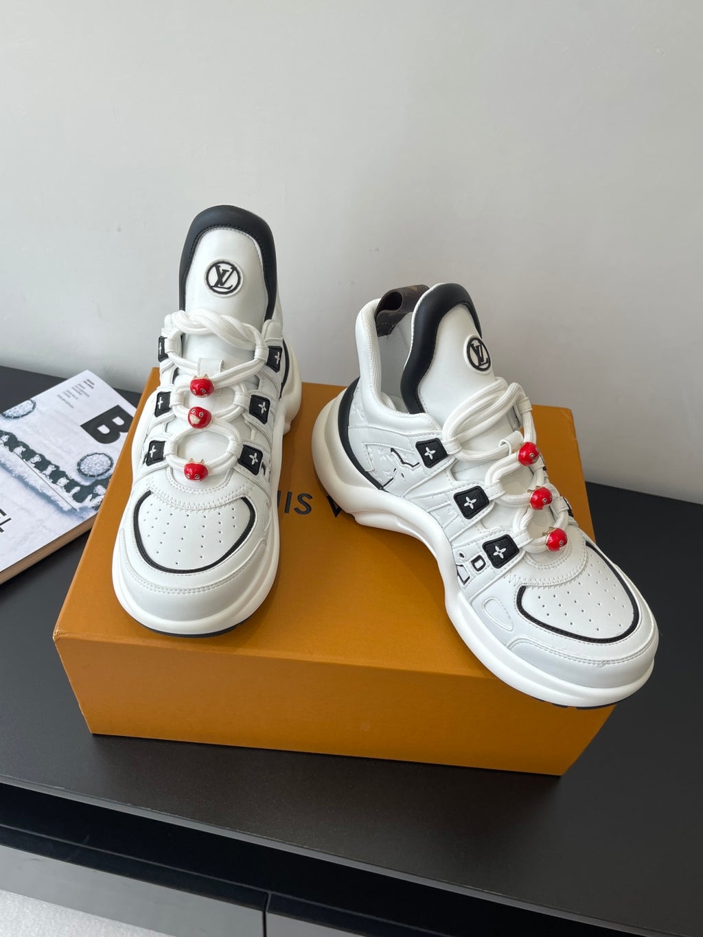 White low-top sneakers with black trim, LV tongue logos, red beaded lace accents