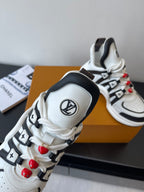 White sneaker close-up with black trim, LV badge, and red beaded lace charms