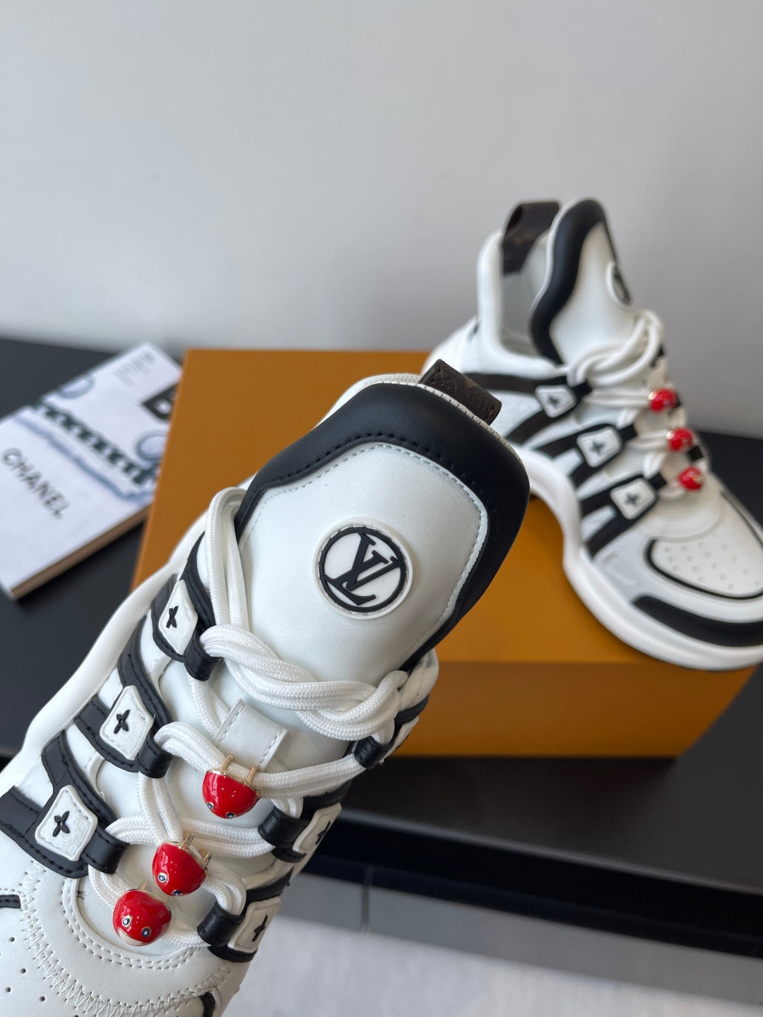 White sneaker close-up with black trim, LV badge, and red beaded lace charms