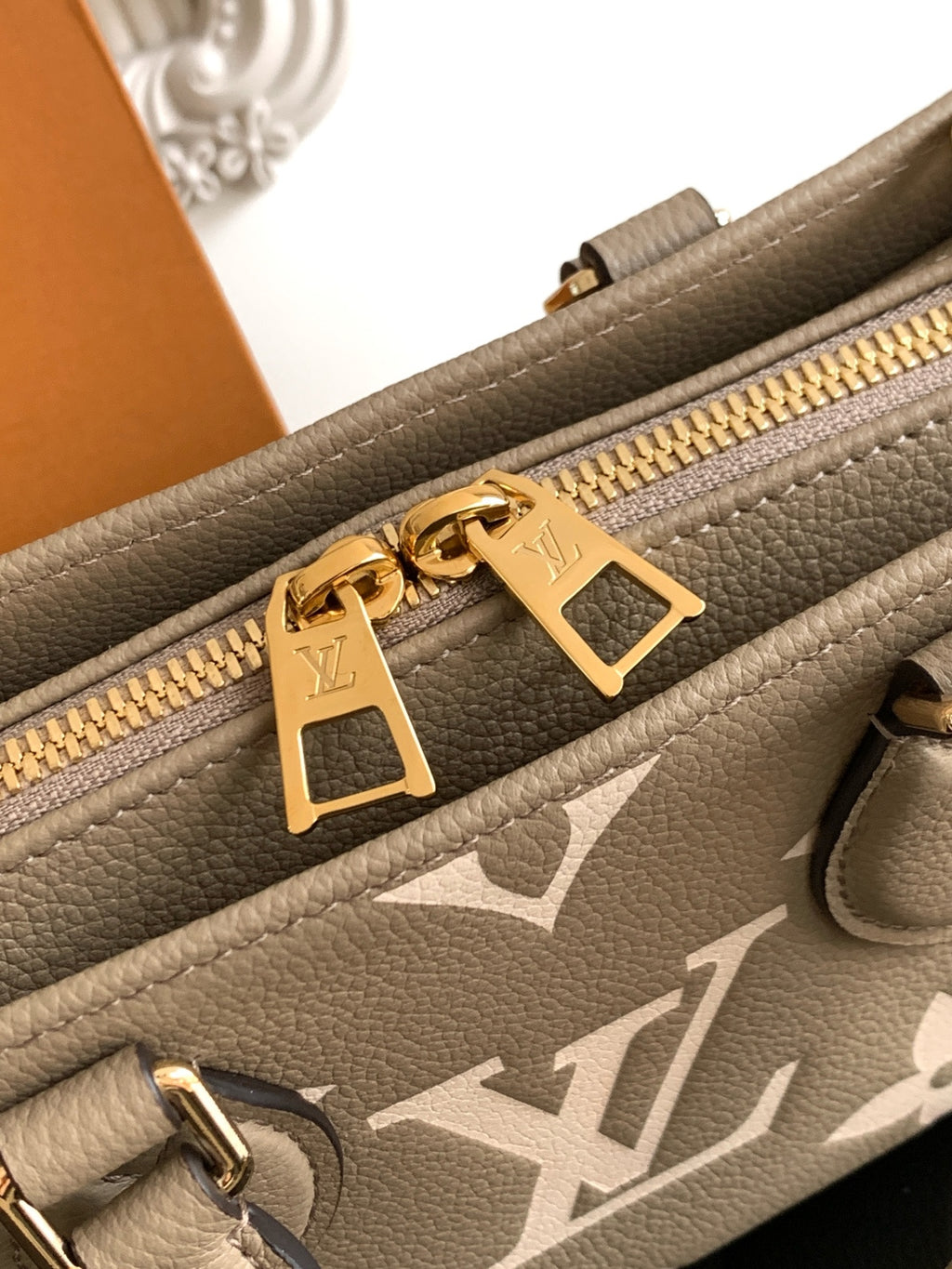 Gray monogram handbag close-up: gold zipper pull and logo plate on leather