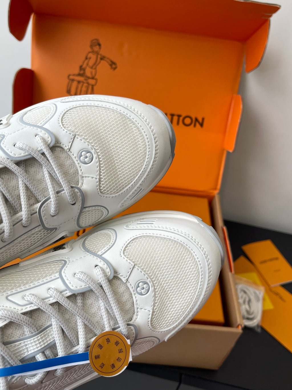 Close-up of white sneakers with textured panels and lace-up eyelets, in orange box.