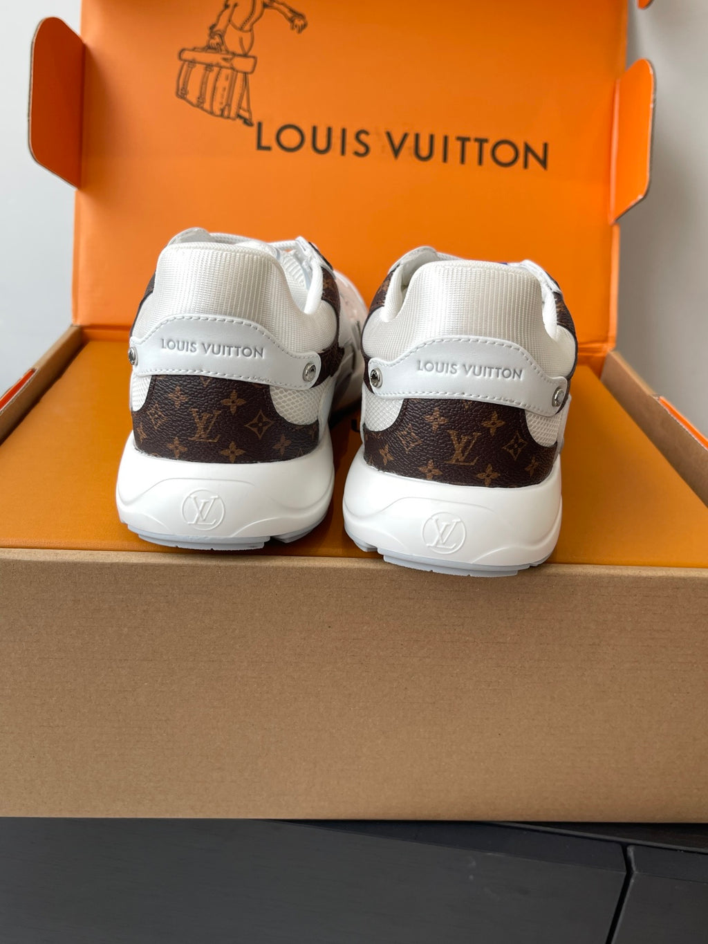Back view of two white sneakers with brown logo heel accents, inside orange shoe box.